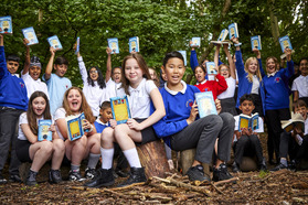 Large group of Year 6 school children situated in a clearing, holding up books with joyful expressions. Two sit on a tree stump in the foreground.
