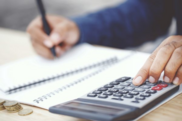 Image of a hand holding a pen and writing on paper along side a calculator 