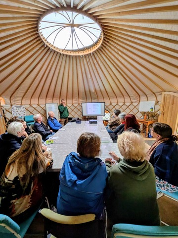People sitting around a table in a yurt with skylight, watching presentation