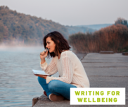 women sat staring at a lake with pen and paper in hand