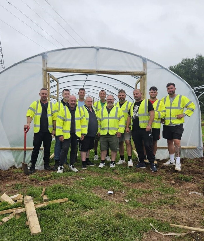 11 volunteers in hi vis jackets standing in front of the poly-tunnel they built