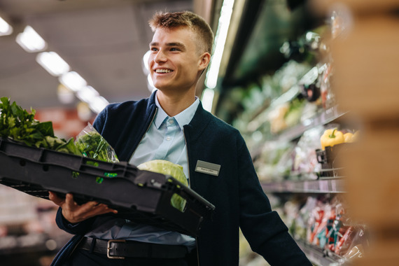 Retail assistant stocking vegetable section whilst greeting out-of-shot customer