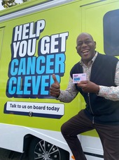 Ainsley Harriott is standing next to a bright green vehicle featuring a large sign that reads "Help You Get Cancer Clever.