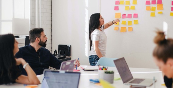 A group of people sitting at a table in a room, with laptops open and yellow and pink post it notes on the wall