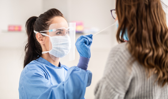 A medic wearing blue PPE, white face mask and a clear visor takes a nose swab from a patient. 