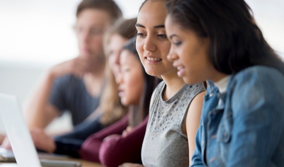 Photo of students sitting at a row of computers collaborating on research. 