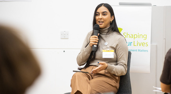 Sukhjeen Kaur from Chronically Brown, a young Asian woman, sitting on a chair holding a microphone, speaking in front of a Shaping Our Lives sign.