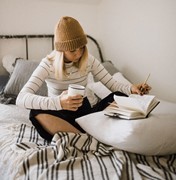 Female student sitting on her bed holding a drink while making notes in a book