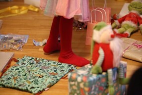 Photo shows various wrapped presents on the floor with a pair of child's legs standing in the middle of them.