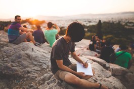 Child sits on rock near beach writing in a notebook