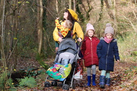 Mother, toddler and 2 young girls walk through an autumnal woodland