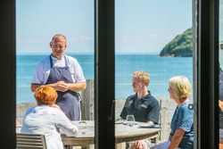 chef talking with customers at a table with seas behind him