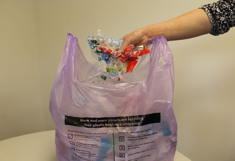 Hand putting flexible plastic packaging into a purple carrier bag