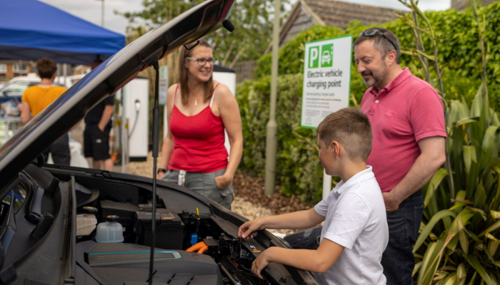 Family stand around an electric car with bonnet up, they're happy and smiling 
