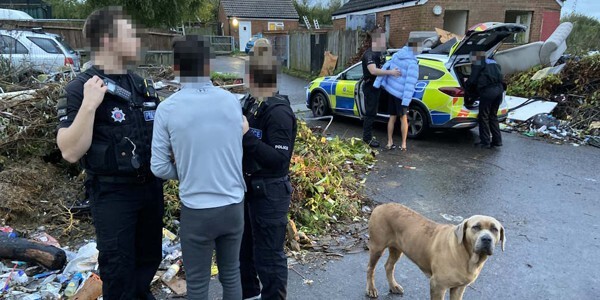 Two suspects are taken to waiting police cars. Another officer is with two more individuals and a dog.