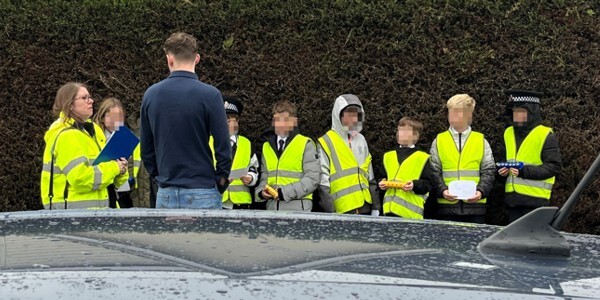 Seven school children wearing high visibility jackets with a police officer and school teacher outside school