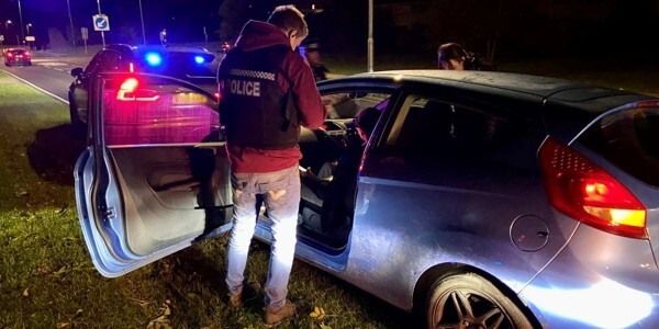 Plain-clothes officer wearing police-marked body armour standing next to a car with the passenger door open at night.