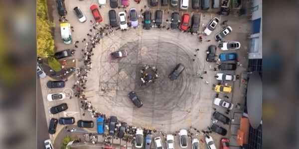Aerial view of a car meet in progress; black tyre skid marks in a circle surrounded by cars and onlookers.