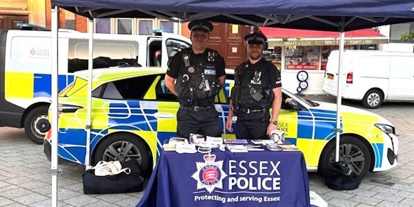 Two officers at an Essex Police tabletop stall outside with police vehicles behind