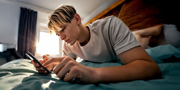 Young man in a T-shirt paying on a bed browsing on a smartphone