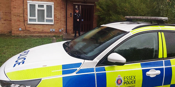 Police Officer stands on door step with marked police car parked in front of vehicle