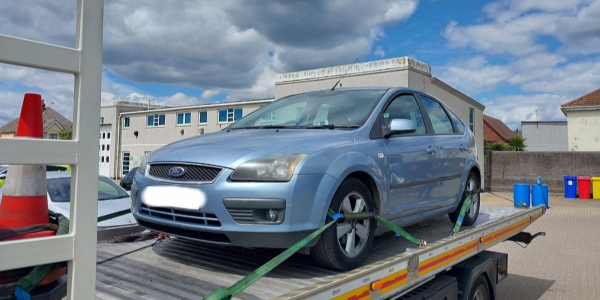 A blue Ford Focus car is loaded onto a flatbed recovery truck, secured with straps, under a cloudy blue sky.