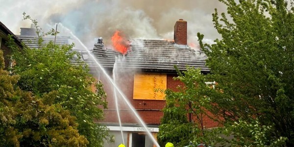 Fire service spraing water with hoses onto a house on fire with significant smoke rising from the roof