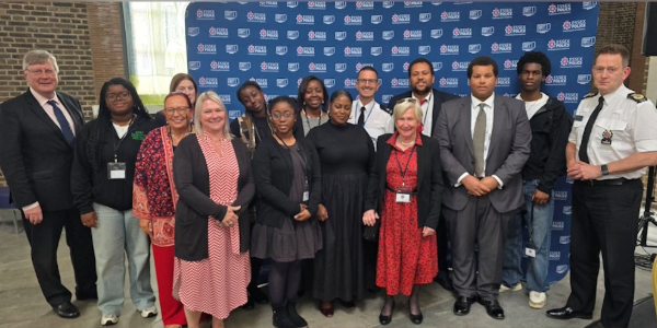 A diverse group of thirteen people posing for a formal photograph in front of an Essex Police branded backdrop.