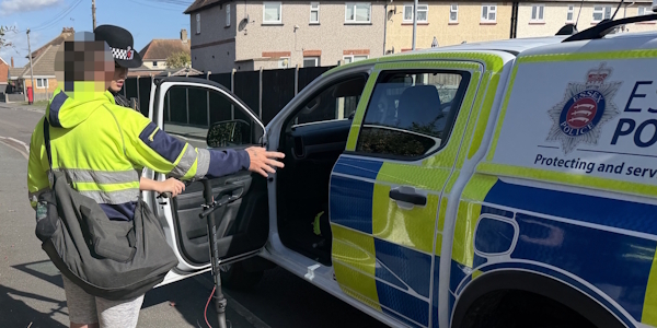 A uniformed Essex Police neighbourhood policing officer speaks with a person next to an e-scooter next to a marked Essex Police vehicle on a street