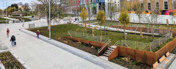 People walk through a modern urban park with young trees, benches, and wide paved paths.