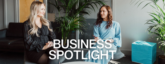Two women sit in a business lounge, talking and smiling, surrounded by green plants. Bold text reads "Business Spotlight."