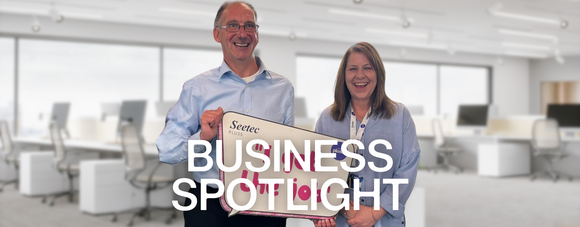 Two people stand side by side in an office, holding a large sign that reads "Seetec Pluss." 