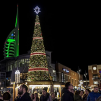 Night time photo of Gunwharf Quays Christmas tree & the Spinnaker Tower lit up green in the background. People are enjoying the Christmas market.