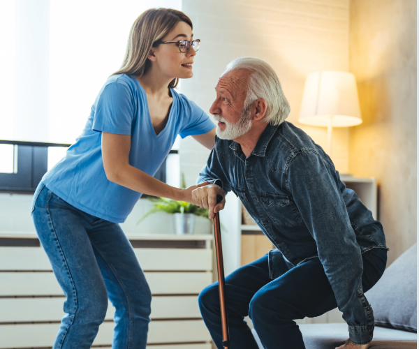 A carer supporting an older adult as they stand up from a chair using a walking stick in a home setting.