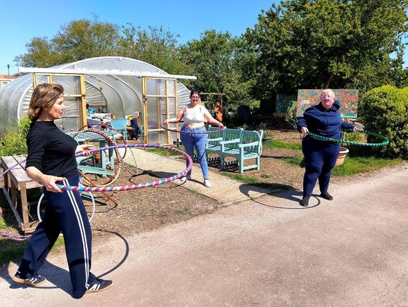 Three people hula hooping outdoors near a polytunnel greenhouse, with benches and trees in a sunny community garden