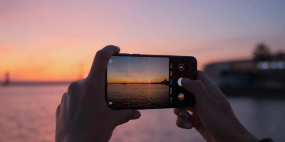 Hands holding a smartphone photographing a sunset over water, with the shoreline and sky reflected on the phone screen
