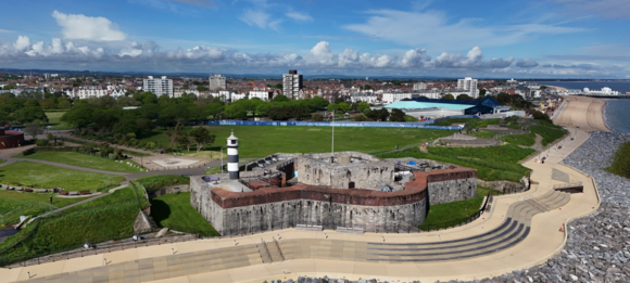 Aerial view of Southsea seafront showing a historic stone fort, lighthouse, green park, promenade and beach under blue sky with clouds