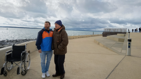 Two people standing together on a seafront promenade beside a wheelchair, with the sea, rocks and dramatic cloudy sky behind them