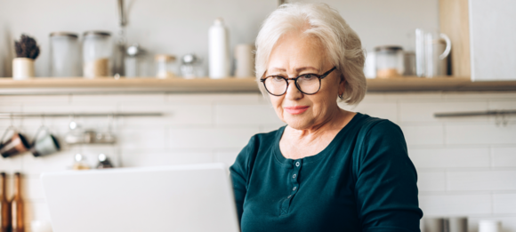 Person wearing glasses using a laptop in a bright kitchen, with shelves, jars and cookware visible in the background