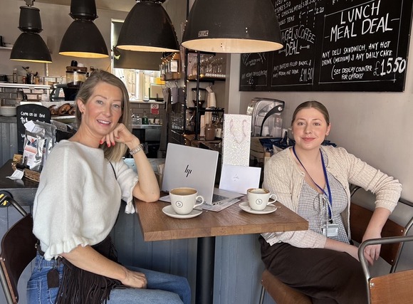 Two people seated at a café table with coffee cups and a laptop, beneath hanging lights and menu boards