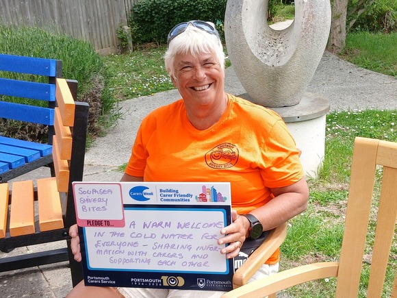 Person holding a Carers Week pledge board outdoors, pledging a warm welcome for carers, seated beside colourful benches and a sculpture