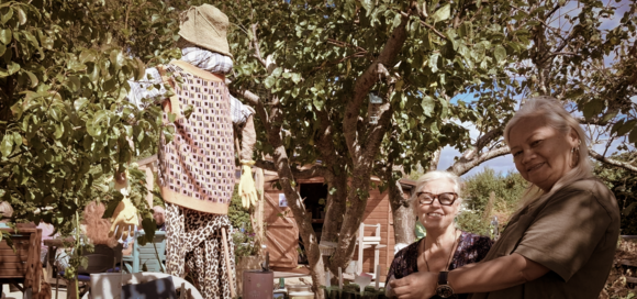 Two carers sitting in a community garden beneath fruit trees, with a scarecrow, shed and allotment plants in the background