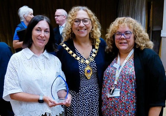 Three people posing with an award at a formal event, including one person wearing a ceremonial chain of office.