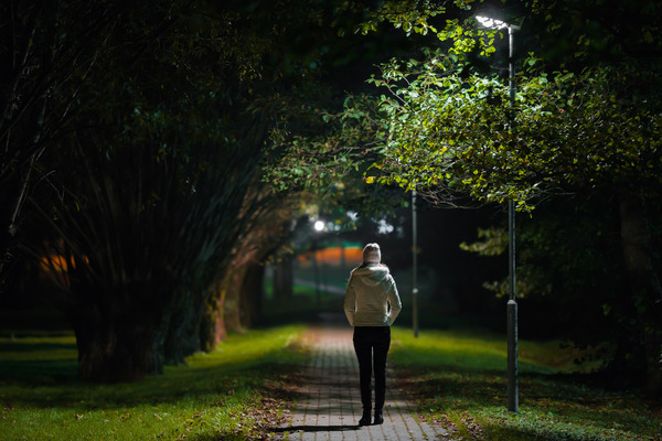 One young alone woman in white jacket walking on sidewalk through alley of trees under lamp light in autumn night