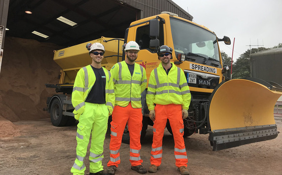 Three male Highways operators standing in front of a gritting lorry with a pile of salt grit in the background.