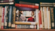 Two people talking behind stacked books in a library.