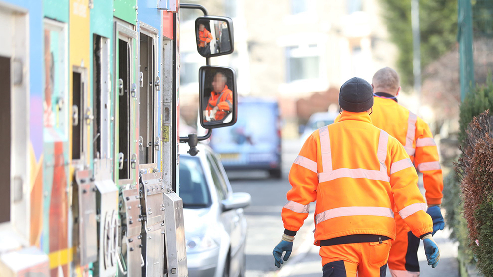 Waste collectors walking up a street with a recycling lorry following behind.