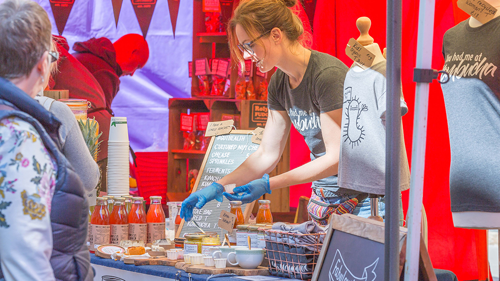 A market stall holder serving sample condiments to a customer.