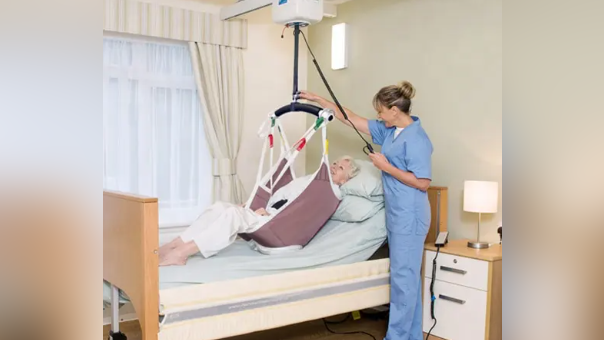 Carer helps elderly woman into bed using a hoist.