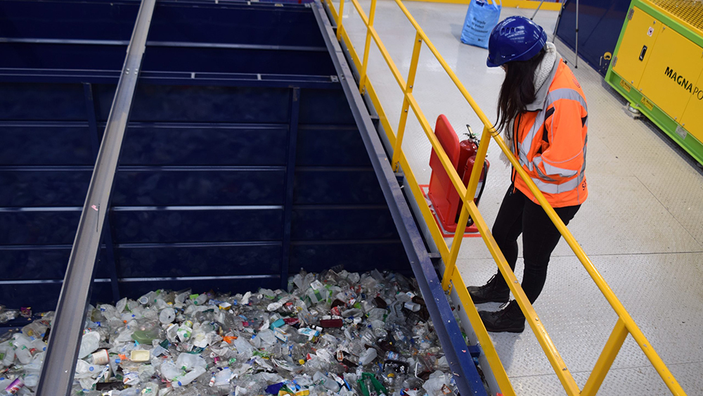 An employee wearing an orange hi-vis jacket and blue hard hat overlooks a sorting bay loaded with plastic.